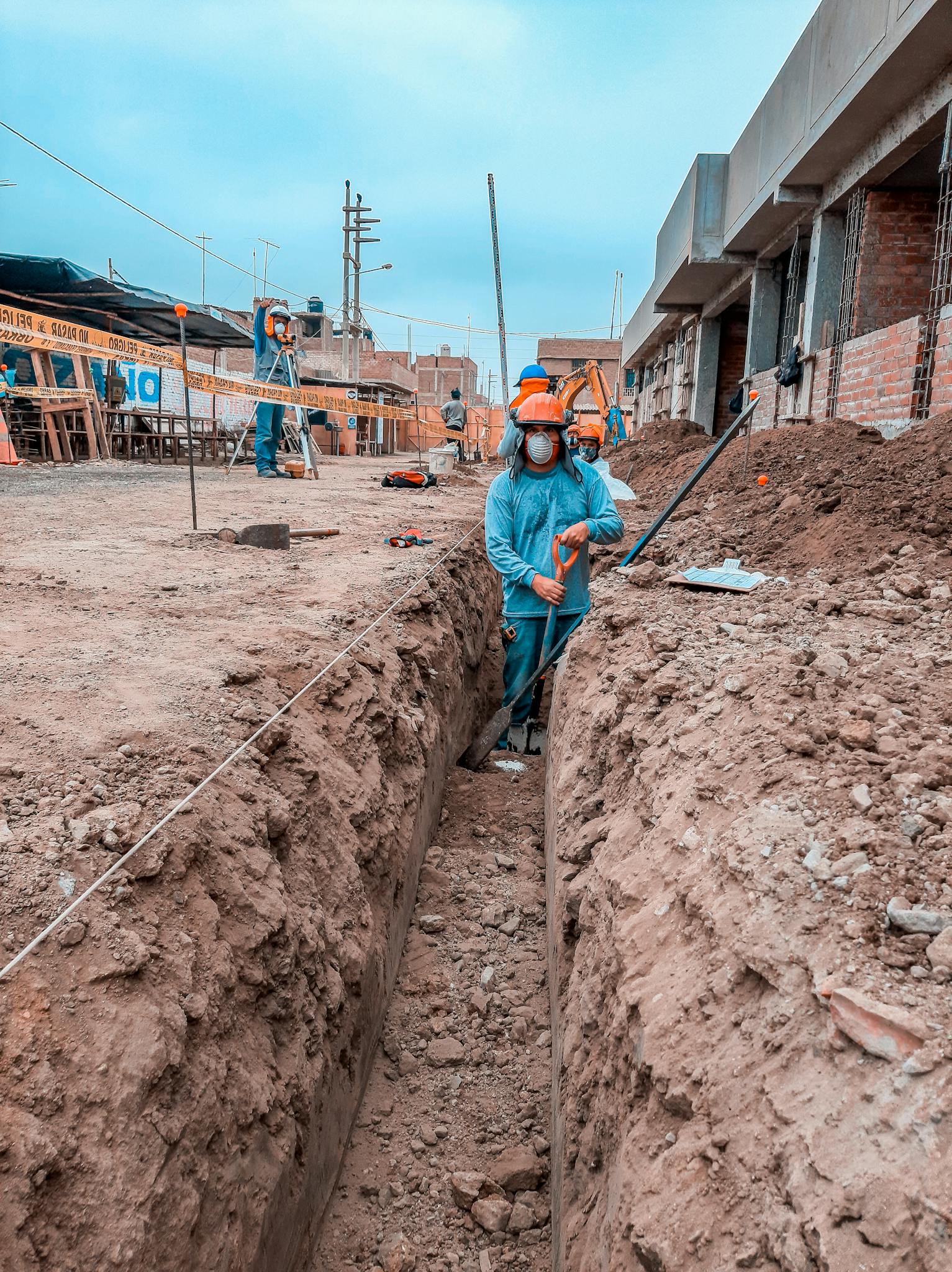 Construction workers in protective gear digging a trench at a site with buildings in the background.
